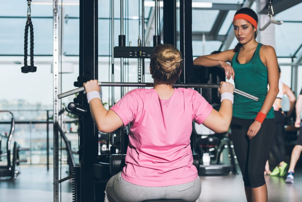Vrouw met overgewicht word begeleid door een personal trainer terwijl ze lat pulldowns doet